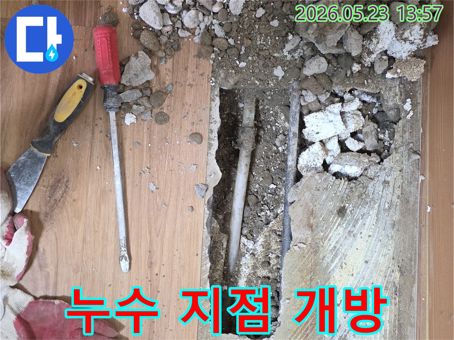 A close-up view of a floor being excavated-revealing a white pipe and scattered debris-with a red-handled screwdriver and a yellow scraper-indicating a precise leak detection process.